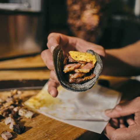 a close up image of hands serving a taco with blue corn tortillas, roasted meat and a piece of pineapple sitting on top