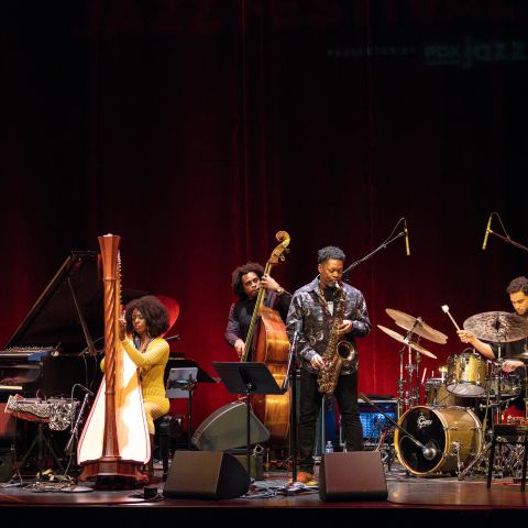 A band of five Jazz musicians performs on stage in front of a red curtain.