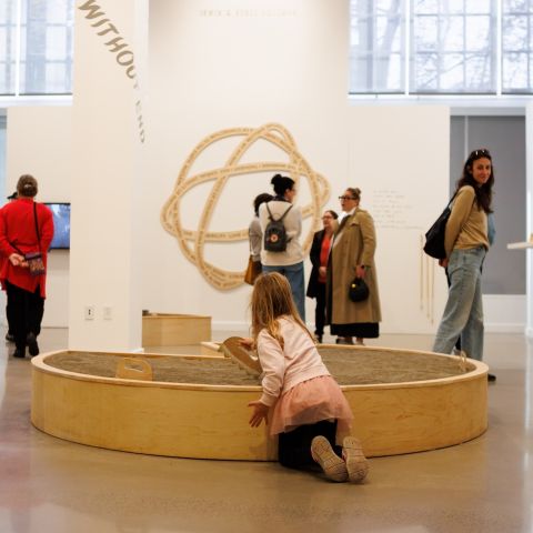 several people browse hung art on the walls of a museum while a child plays in a central sand box