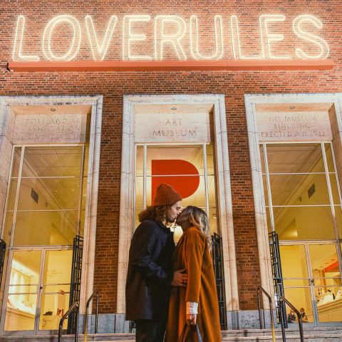 Two people kissing in front of the \"Love Rules\" sign at Portland Art Museum.