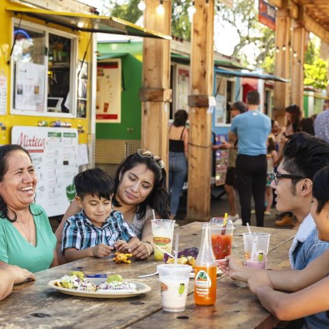 people sitting at a picnic table in front of a line of colorful food carts