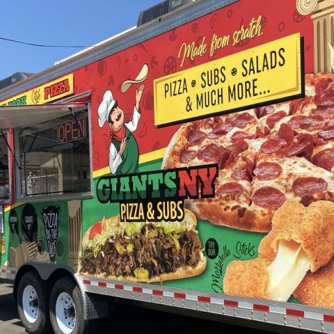 a red and green food cart with a sign advertising pizza and subs beneath clear blue skies