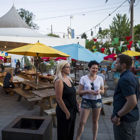 people stand in a food cart pod patio with colorful umbrellas and flags covering picnic tables