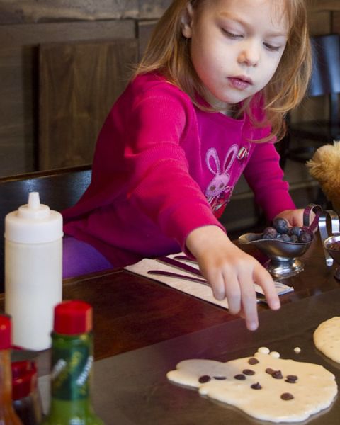 a young girl adds chocolate chips and blueberries to a pancake