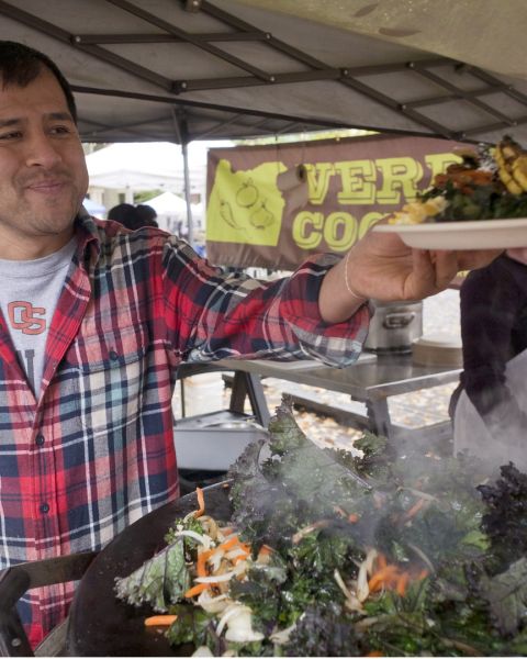 man in an outdoor market handing a plate of food to a customer in front of a sign reading \"Verde Cocina\"