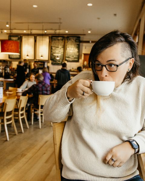 a white person with long hair on one side and a shaved head on the other side, sips from a coffee mug in a warmly lit bakery.