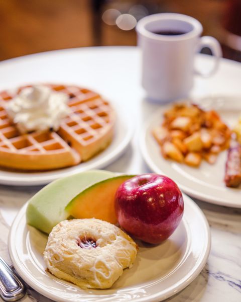 table with traditional breakfast foods, waffle, home fries, sausages, scrambled eggs and fruit with a pastry