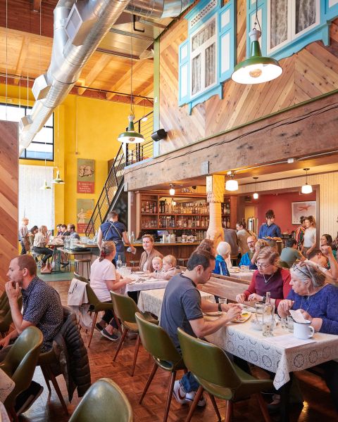 tables full of diners enjoy a meal in a lively, warmly lit restaurant with a bar in the background