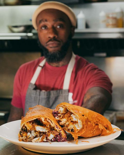 a Black man in a red shirt, yellow beanie and apron presents an overstuffed burrito on a plate