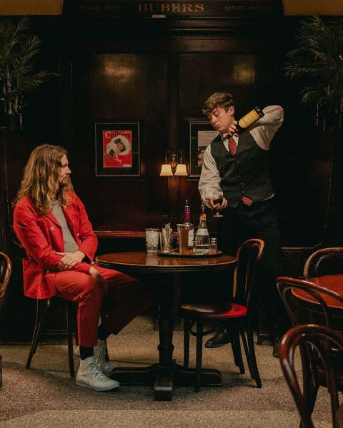 a server in an atmospheric restaurant prepares a cocktail for a man in a red suit seated at a table