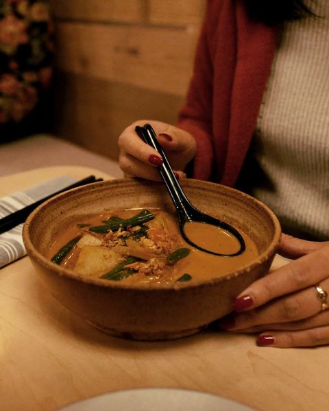 A person sitting at a table holds a ramen spoon in a bowl