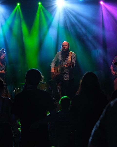 a band plays in front of a crowd under green, blue, and purple stage lights