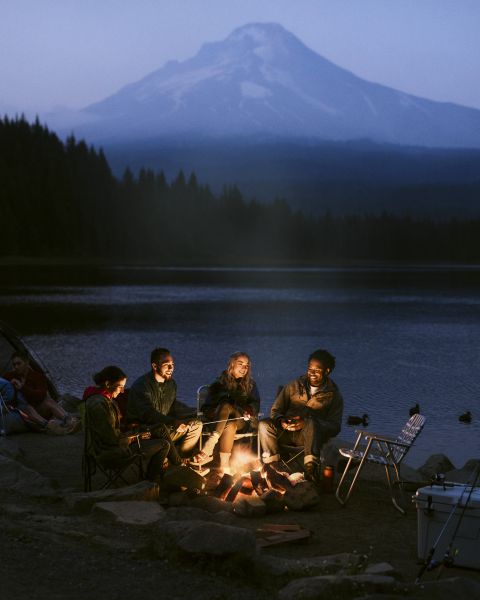 four people illuminated by campfire at a dimly lit campsite, backed by a serene lake and a large mountain obscured by mist