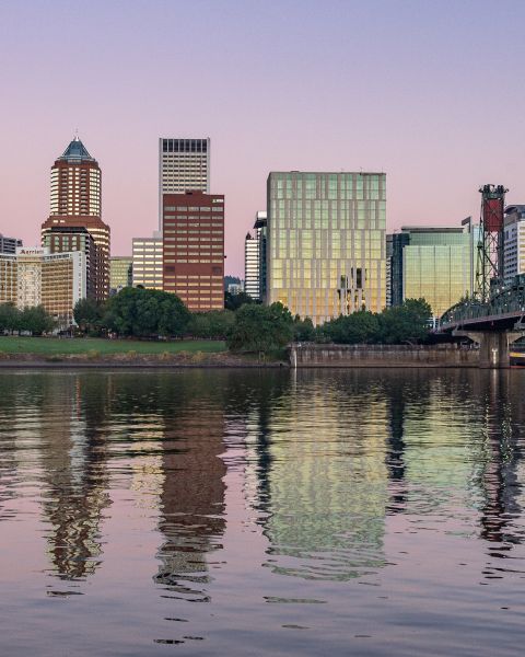 pale pink and purple sky reflect on a river surface with a bridge off to the side and a view of downtown in the background