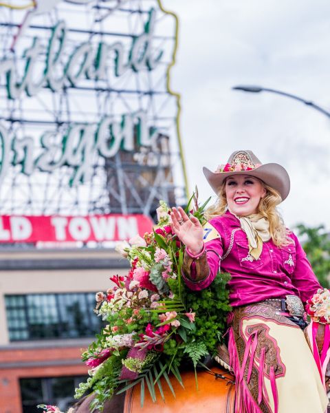 woman in hat and chaps ride a rose decorated horse in a parade with Portland\'s white stag sign in the background