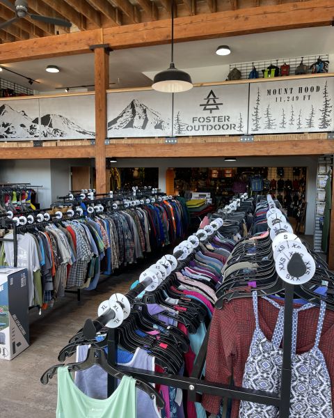 Racks of new and used outdoor clothing sit in a bright space accented by wood beams and the Foster Outdoor mountain scape logo.