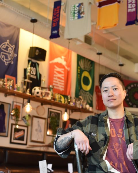 A woman in a plaid button-down and red t-shirt sits at a bar in front of a wall of women\'s sports memorabilia.