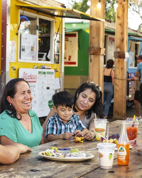 people sitting at a picnic table in front of a line of colorful food carts