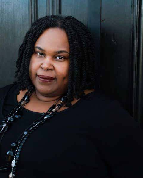 A Black woman with dark mid-length twists, a beaded necklace and a black shirt subtly smiles into the camera.