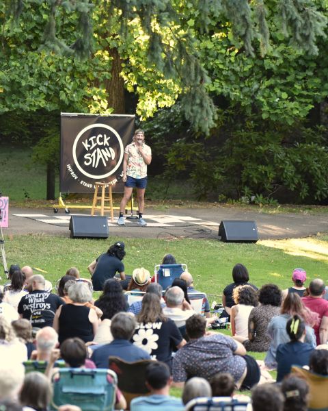 Blond comedian on stage performing in front of large crowd sitting in a park