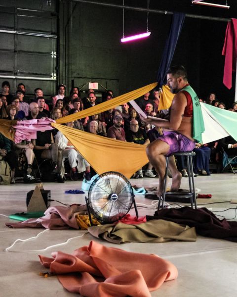 a person sits on a stool in front of an audience with colorful clothing strewn about the stage