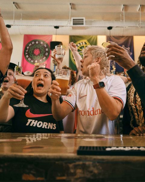 a crowd of people cheer while raising glasses in front of a bar while watching a game