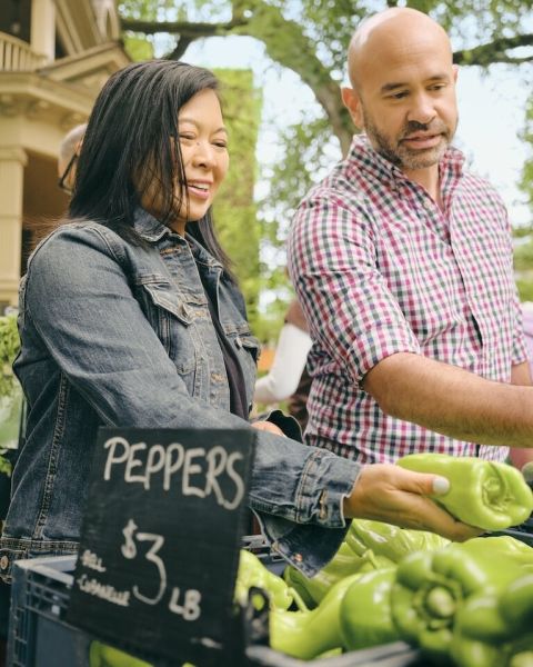 two people peruse fresh produce like peppers and zucchini outside in bins with signs saying what each bin contains and how much they are a pound
