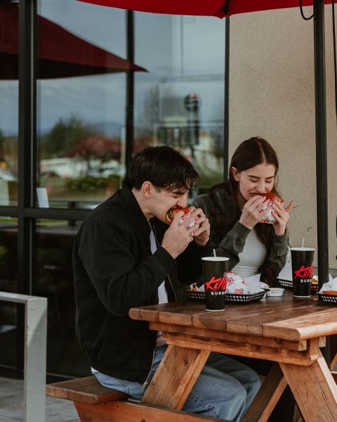 A couple sits at a picnic table eating burgers.