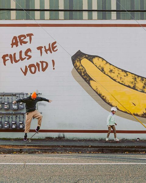 A man and a child skateboard on a sidewalk in front of a mural of a yellow banana with text reading. \"Art fills the void!\"