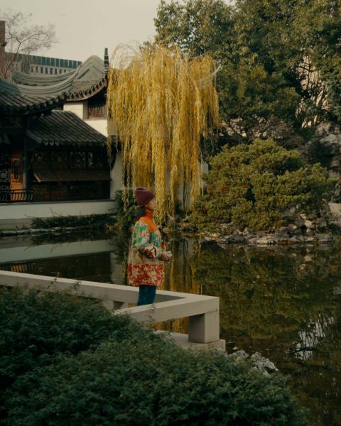 A woman holds a camera to her eyes in a traditional Chinese garden with a pond, pavilion and trees.