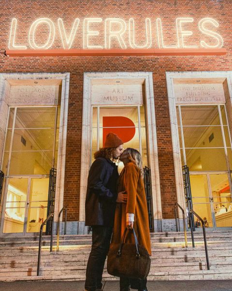 Two people kissing in front of the \"Love Rules\" sign at Portland Art Museum.