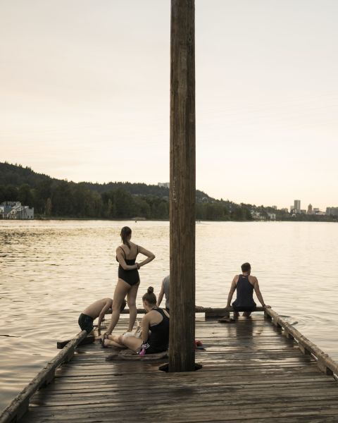 a wooden dock in a river — with swimmers standing, sitting or climbing out of the water — looks toward a city skyline in the fading sunlight