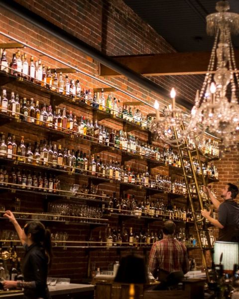 several bartenders peruse shelves housing a vast collection of whiskey bottles