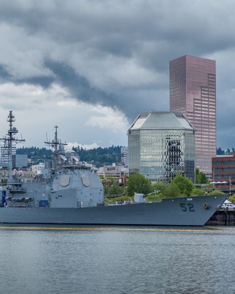 large warship docked at Waterfront Park in Portland with dramatic grey clouds over buildings in downtown