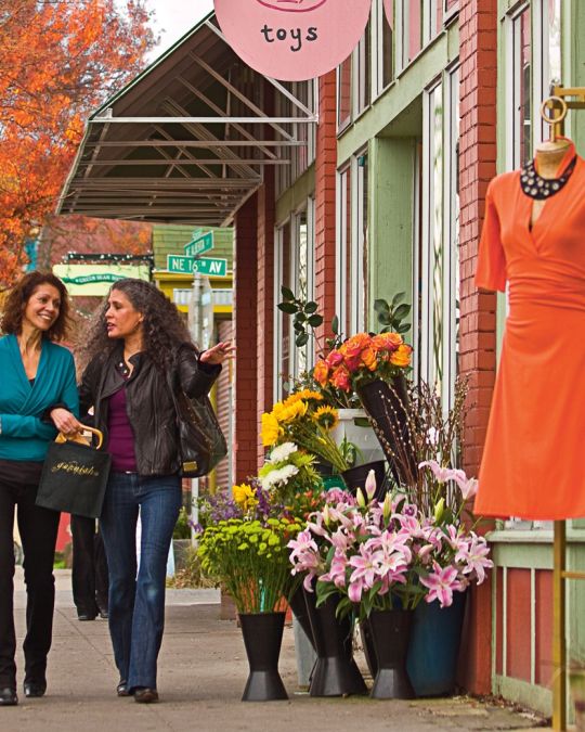 Two shoppers walking down the sidewalk in front of shops displaying flowers and clothing on Northeast Alberta Street