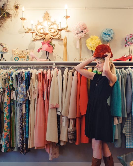 a woman trying on a hat in front of racks of vintage clothing