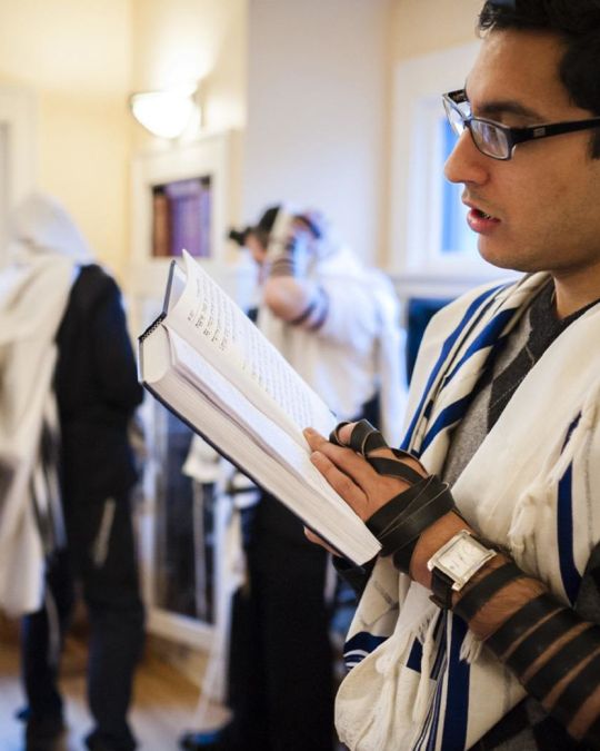 a man wearing a shroud and yamaka, reading the Torah