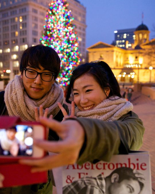 A couple poses for a selfie in front of a Christmas tree and a historic building