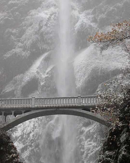 a towering waterfall behind a stone bridge surrounded by snow and ice