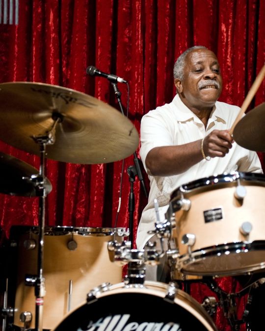 a man playing drums on stage in front of a red curtain