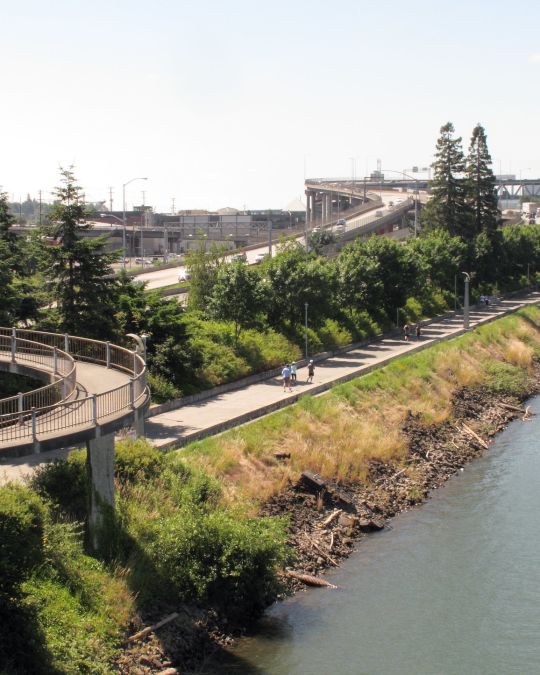 Three people use a riverside path flanked by a highway