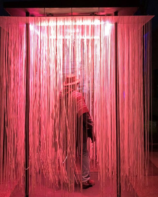a person walks through a light installation containing long strands of lights hanging from a panel overhead