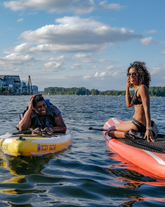 Two women on paddleboards in a river with houseboats in the distance