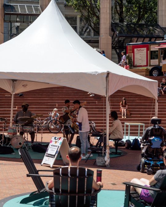 white tent with band surrounded by people in chairs, people sitting on steps in an urban courtyard