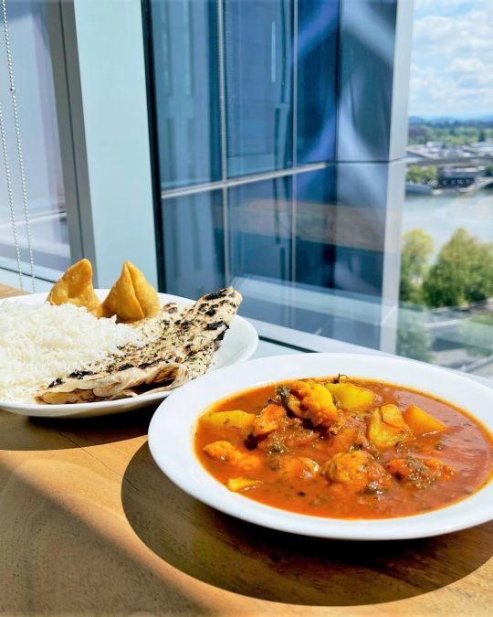 A white plate of aloo gobi sits next to a platter of rice, samosas and naan on a wooden table in front of a large window.