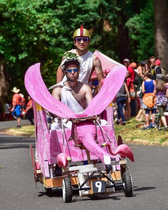 two people wearing sunglasses and togas ride a pink elephant-shaped soapbox derby cart