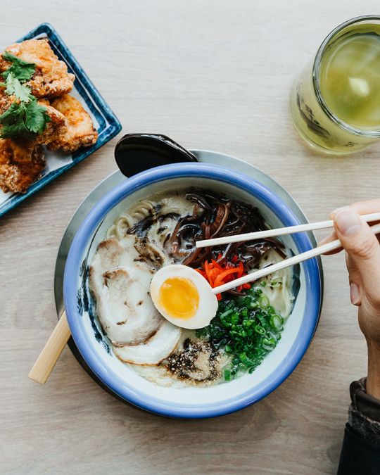 a hand holding chopsticks hovers over a bowl of ramen. a mug of green tea and a plate of fried chicken are next to the ramen bowl on a table.