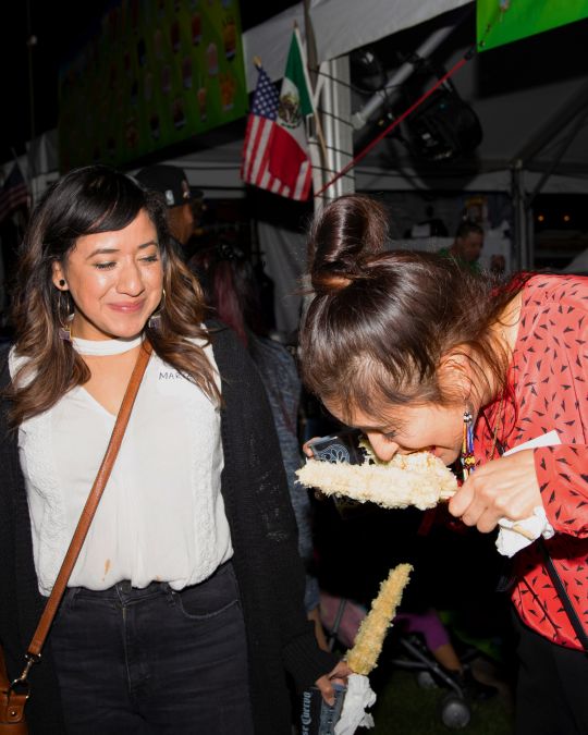 two people standing next to a food cart, one eating an ear of corn