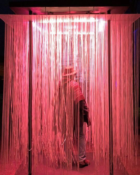 a person walks through a light installation containing long strands of lights hanging from a panel overhead