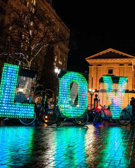a night image of the word LOVE spelled out in large letters made of hundreds of small bulbs attached to bicycles in front of a large brick building